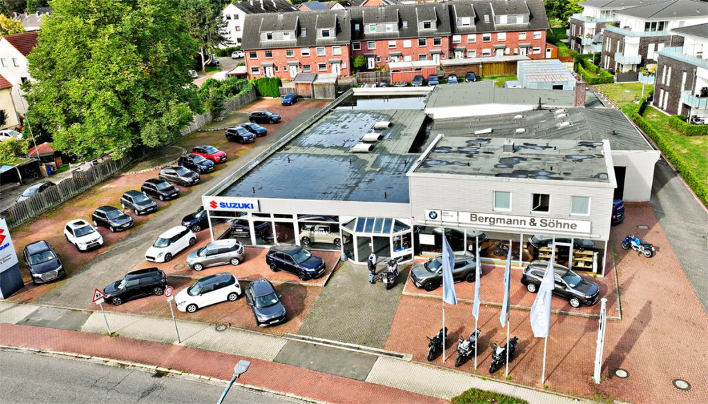 Luftaufnahme eines Autohauses mit Suzuki- und BMW-Fahrzeugen, mehreren abgestellten Autos und Motorrädern vor einem Gebäude in einem Wohngebiet.